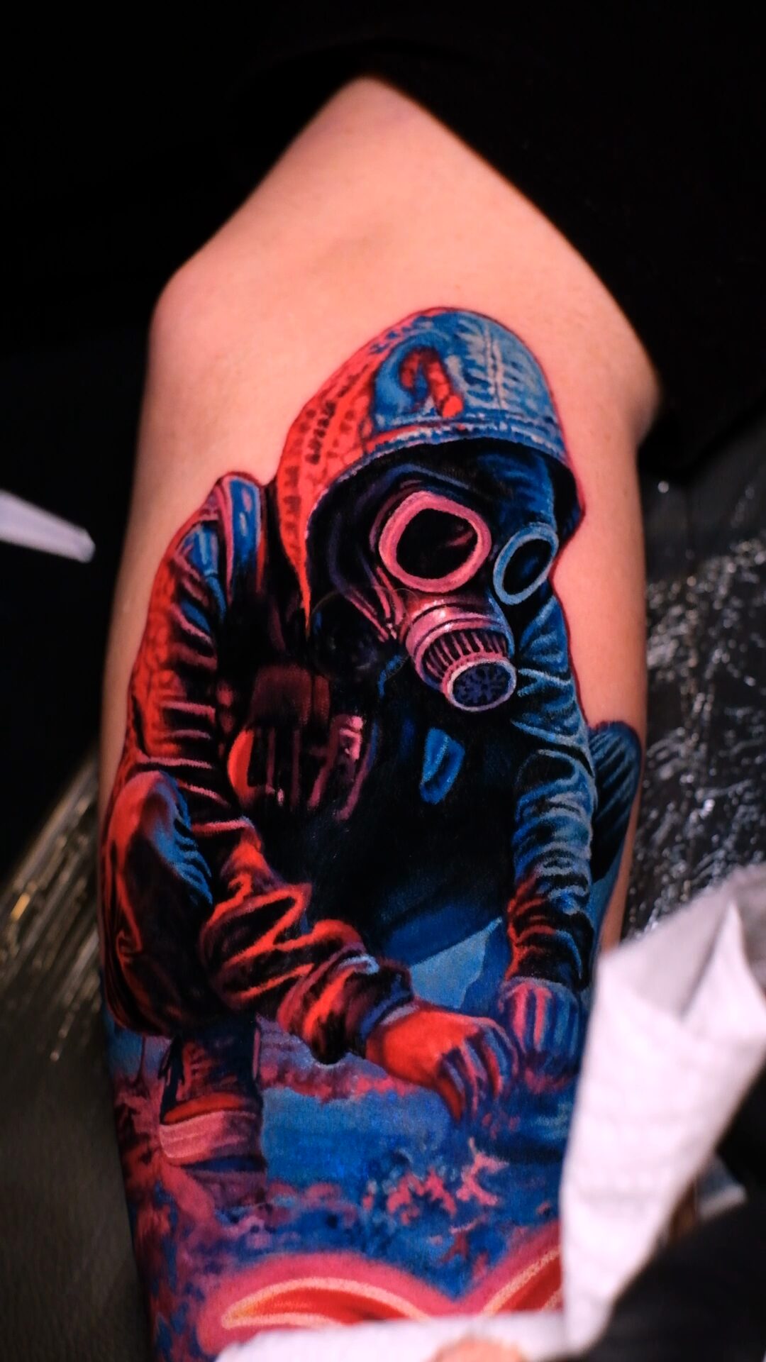 Nahaufnahme: Gasmaske mit rosa leuchtenden Gläsern und Filter, Aufnäher auf der Jacke, Hände auf den Knien, Schuhe – Tattoo auf dem Oberarm.